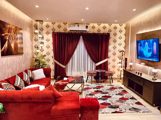 Open living area with crimson curtains, printed rug, TV unit with LED backlight, mosaic floor lamps and split AC.